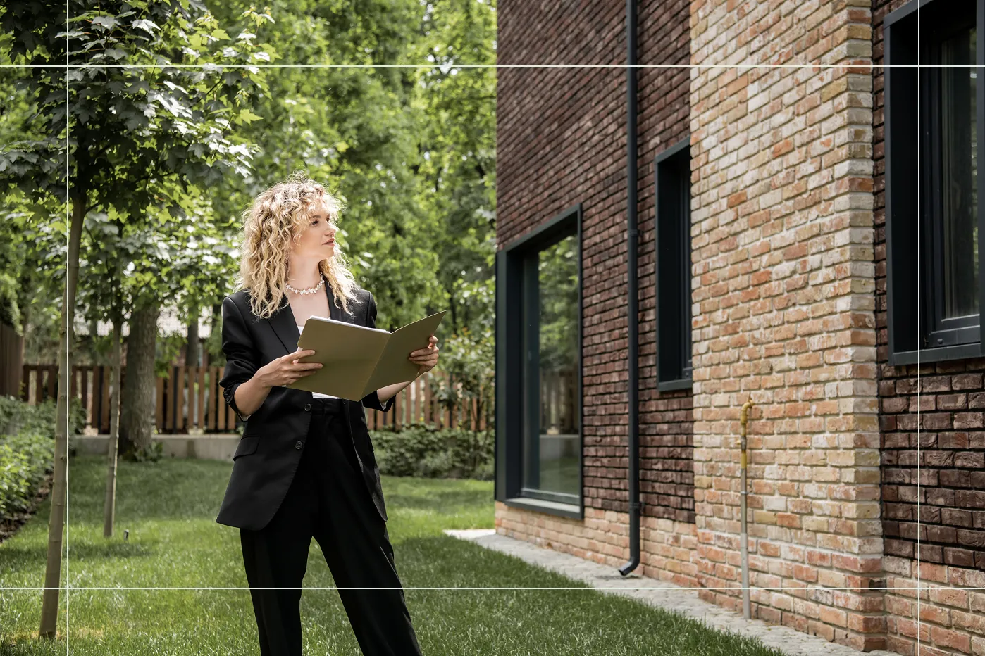 Frau in schwarzem Anzug steht mit geöffnetem Ordner vor einem Backsteingebäude im Garten