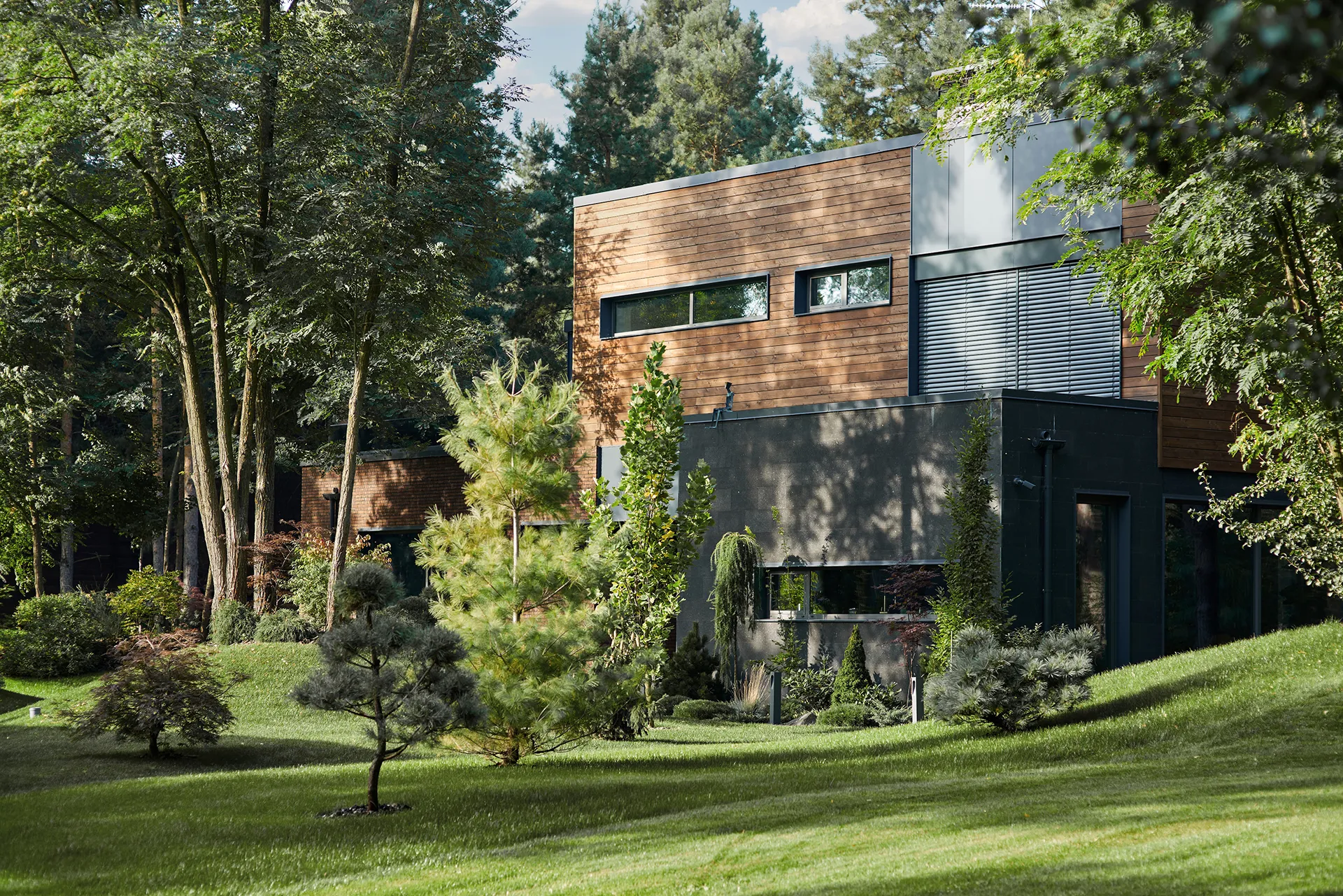 Modernes zweistöckiges Haus mit Holz- und Betonfassade, umgeben von Bäumen und gepflegtem Rasen