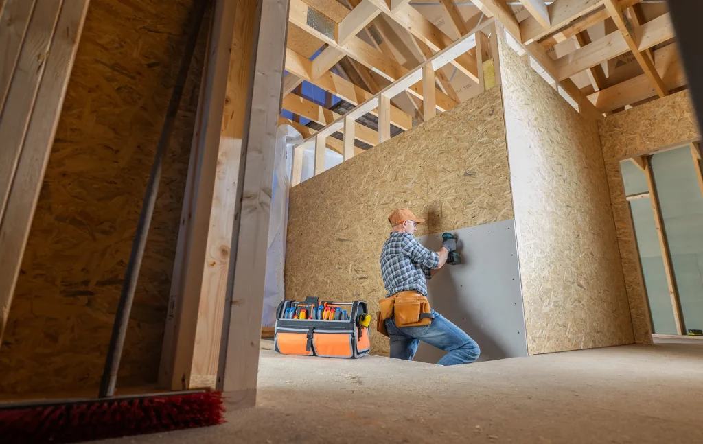 Handwerker mit Kappe und kariertem Hemd befestigt Gipskartonplatte an Wand im Rohbau eines Hauses